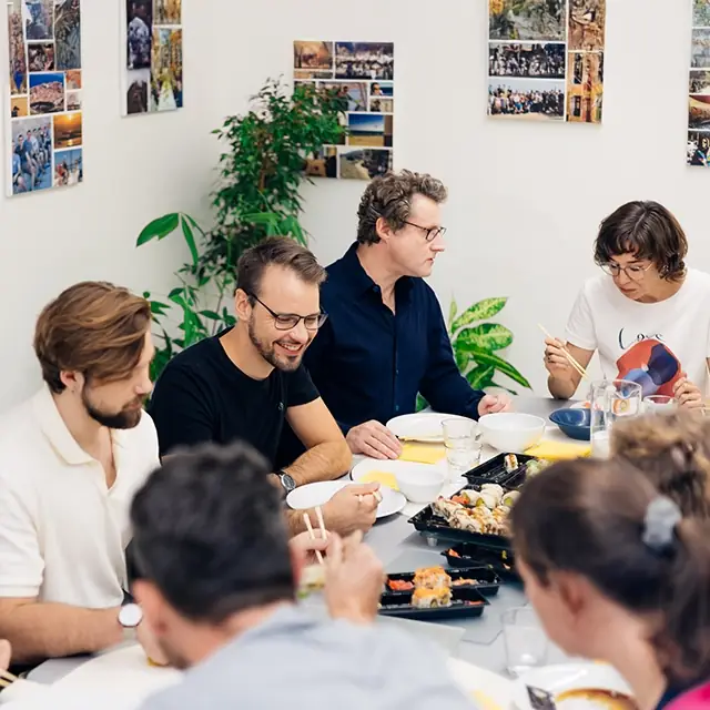 Mehrere Kolleginnen und Kollegen sitzen gemeinsam an einem Tisch und essen Sushi, während sie sich angeregt unterhalten. Im Vordergrund sind Philipp Zelenka, Daniel Winzer, Gereon Friederes und Karin Graf zu sehen. Im Hintergrund sind grüne Pflanzen und Urlaubsfotos an der Wand zu sehen.