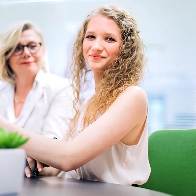 Sophie Buchta sitzt am einem Tisch und wird dabei von Britta Veser bei ihrer Arbeit unterstützt.