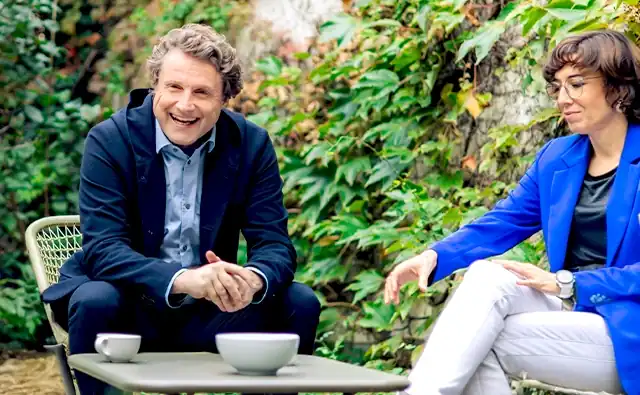 Gereon Friederes sitzt lächelnd in einem Gartenstuhl und führt ein Gespräch, während Karin Graf in blauem Blazer ihm gegenübersitzt und konzentriert mit einer Geste kommuniziert, zwischen ihnen stehen Kaffeetassen auf einem Tisch.