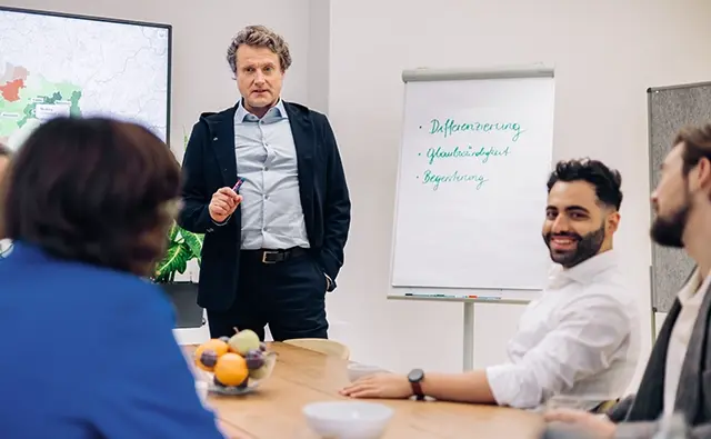 Gereon Friederes steht vor einem Flipchart und spricht zu einer Gruppe von Menschen in einem Besprechungsraum. Auf dem Flipchart stehen die Begriffe Differenzierung, Glaubwürdigkeit und Begeisterung.