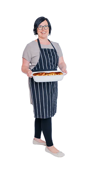 Mag. Anita Salamon trägt eine gestreifte Schürze und hält stolz ein frisch gebackenes Auflaufgericht in den Händen – herzlich, genussfreudig und mit Freude am Kochen.
