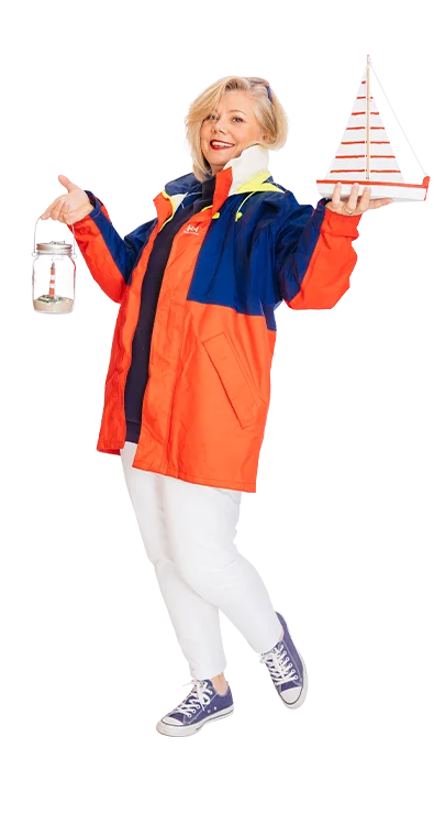 Dipl.-Kff. Britta Veser trägt eine farbenfrohe Segeljacke, hält ein Miniatursegelboot in der einen Hand und eine Laterne mit einem Segelschiff darin in der anderen - sie liebt das Meer und maritimes Flair.