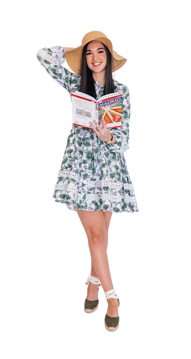 Giselle Kaufmann, MSc trägt ein geblümtes Sommerkleid und einen Sonnenhut, während sie mit einem Madrid-Reiseführer lächelnd Fernweh weckt.