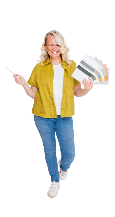 Mag. Kerstin Hofer steht mit Farbfächer und Pinsel in der Hand und lächelt begeistert in die Kamera – kreativ, lebensfroh und voller Gestaltungslust.