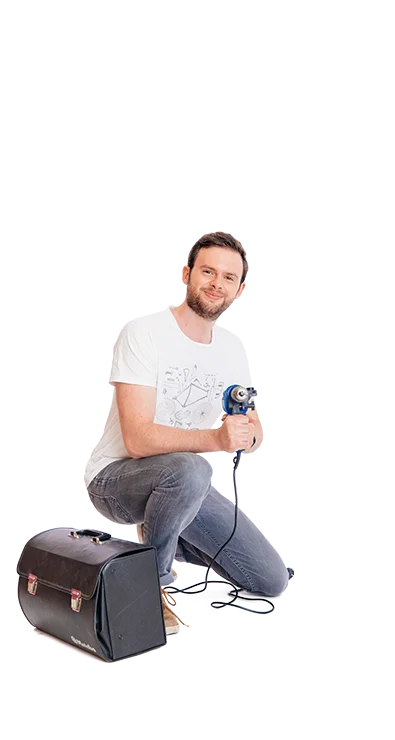 Lucas Pum, MA hockt mit Bohrmaschine in der Hand neben einer Tasche, trägt Jeans und T-Shirt - technikbegeistert, kreativ und fokussiert.