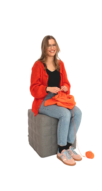 Polina Yaroshchuk, BA MA sitzt mit oranger Strickjacke und Wolle auf einem Hocker, das Strickzeug in der Hand - kreativ, konzentriert und handwerklich geschickt.