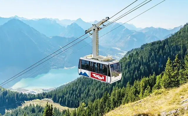 Eine Seilbahn mit Tirol-Branding beim der Bergfahrt - mit Blick auf einen türkisfarbenen Bergsee und bewaldete Alpen.