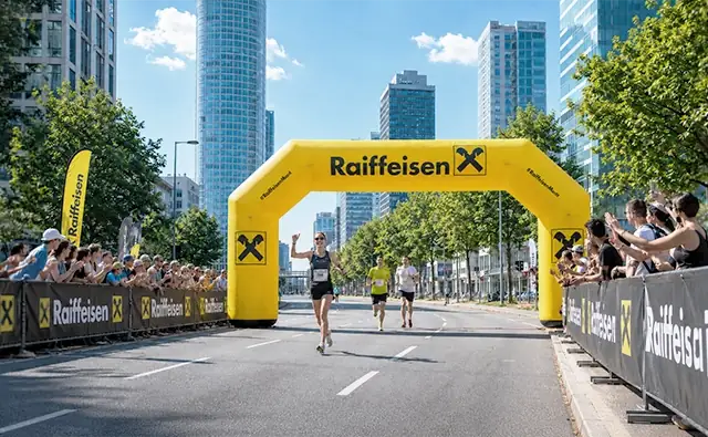 Läuferinnen und Läufer starten bei einem Stadtmarathon unter einem gelben Raiffeisen-Bank-Torbogen.